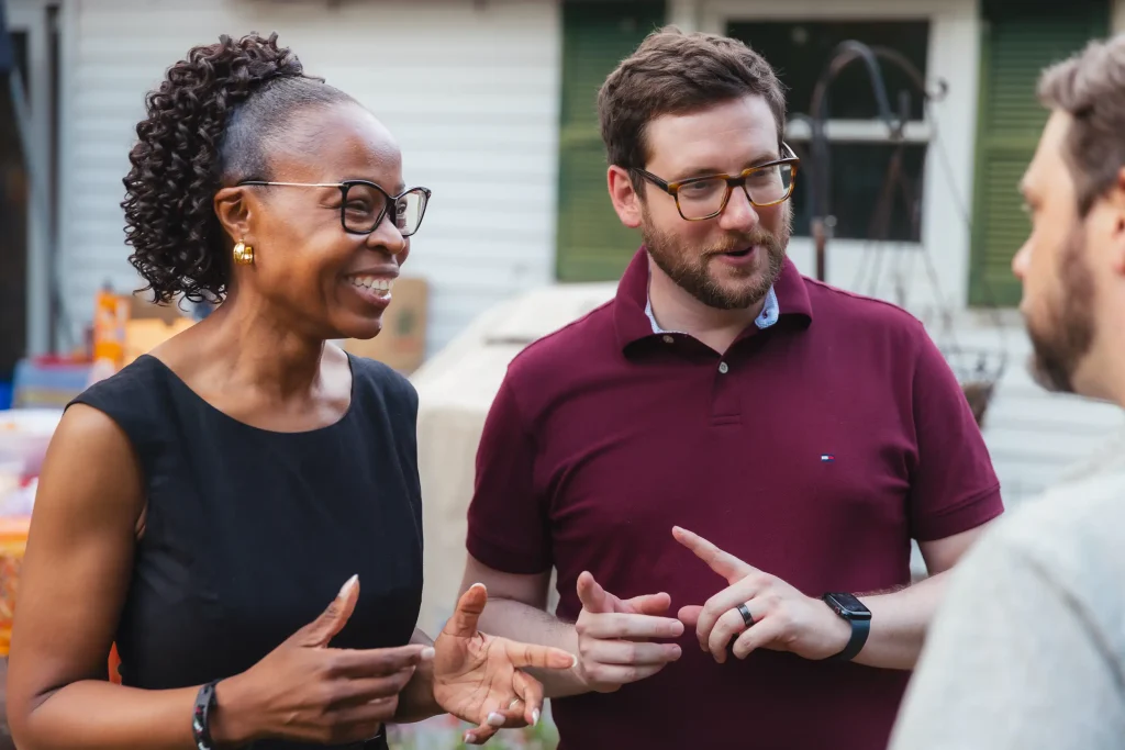 A female politician and a male politician stand next to each other smiling while speaking with a constituent.