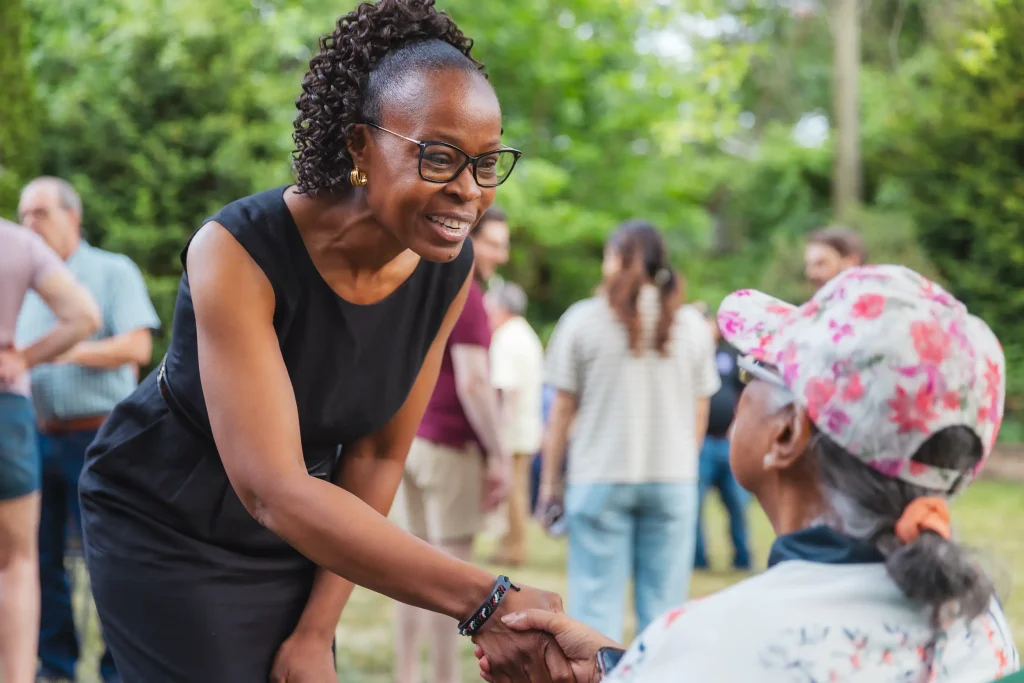 A politician in a black dress shakes the hand of a seated constituent at an outdoor social gathering.
