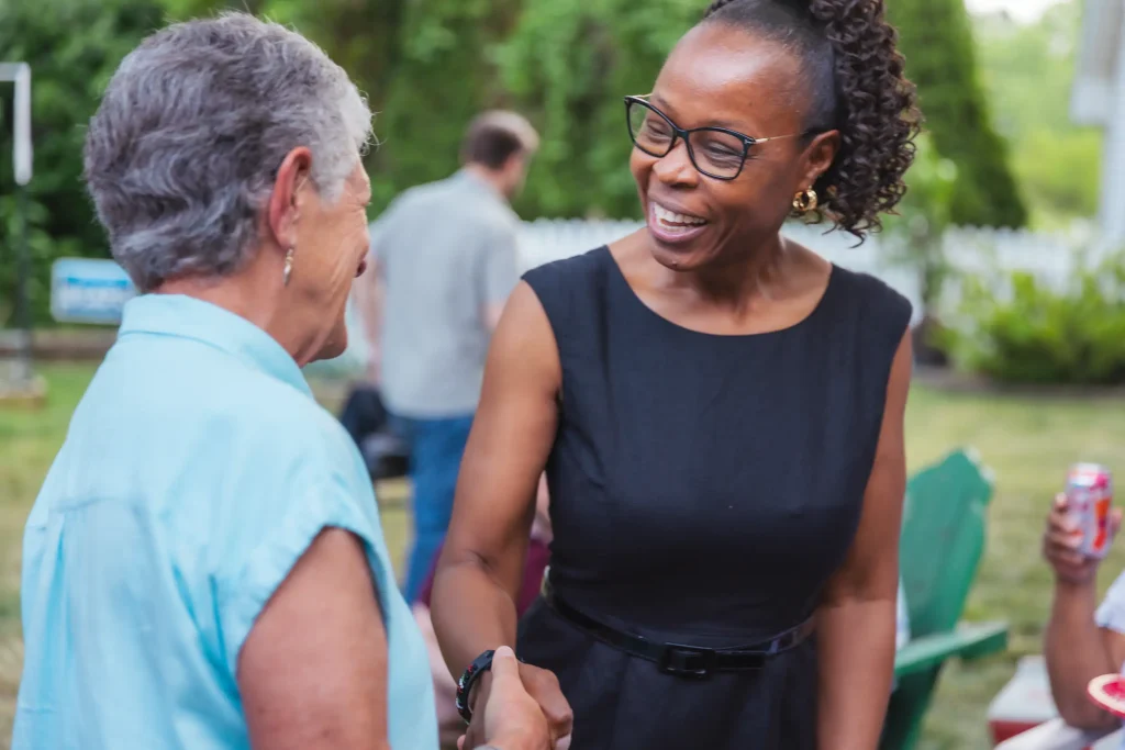 A politician in a black dress smiles and shakes hands with a constituent in a blue shirt.
